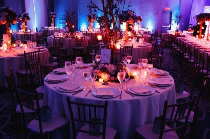 alt= A large, elaborately decorated banquet hall with red lighting, round tables set for dinner, and elegant chandeliers hanging from the ceiling.